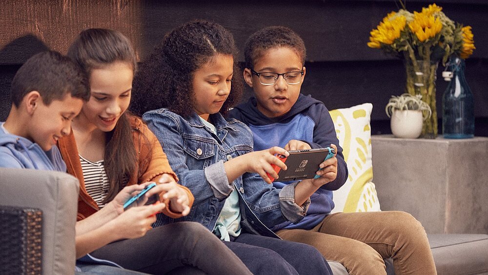 Czworo dzieci siedzi na kanapie, trzymając w rękach kontrolery do konsoli Nintendo Switch. W tle widoczne są żółte kwiaty w wazonie i poduszka z wzorem.