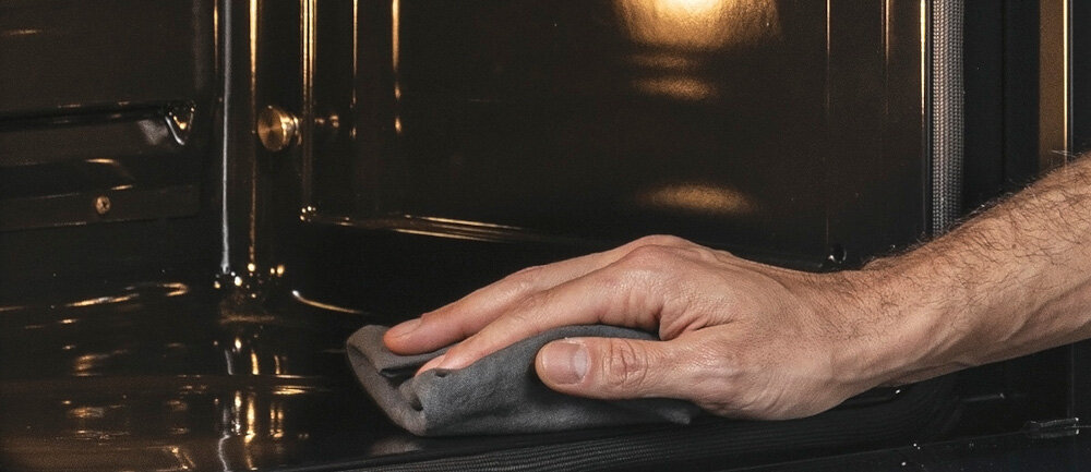 Ręka trzymająca szmatkę przeciera wnętrze piekarnika o metalicznym wykończeniu.