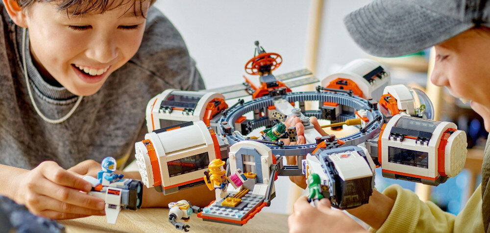 Dwoje dzieci bawi się zestawem LEGO przedstawiającym modułową stację kosmiczną z różnymi elementami i figurkami astronautów.