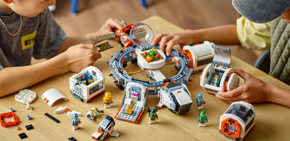 Na stole znajduje się zestaw LEGO przedstawiający modułową stację kosmiczną z różnymi elementami i figurkami. Dwie osoby składają zestaw, manipulując jego częściami.