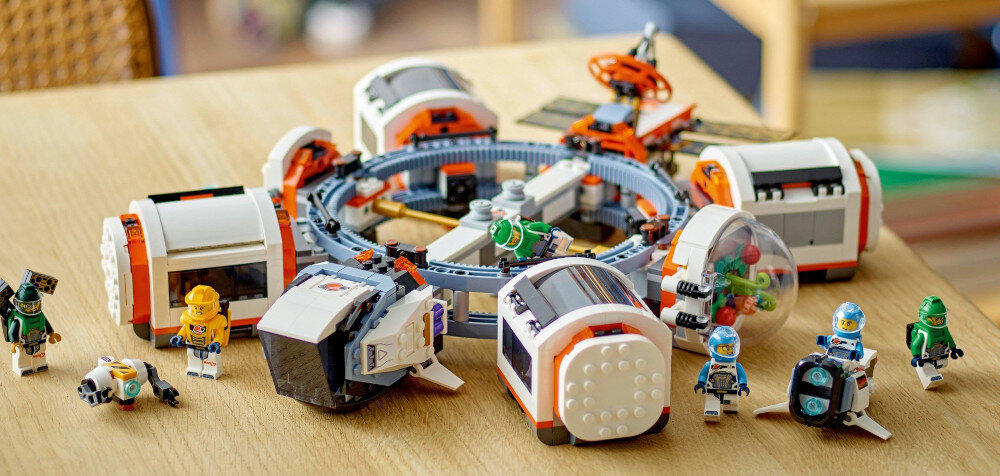 Na drewnianym stole znajduje się zestaw klocków LEGO przedstawiający modułową stację kosmiczną z kilkoma figurkami astronautów. Elementy zestawu są w kolorach białym, pomarańczowym i szarym.