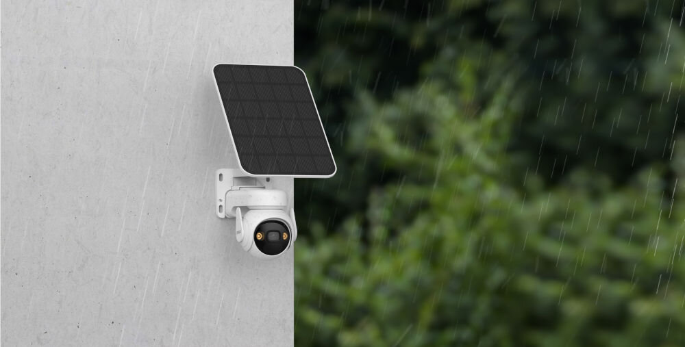 Na ścianie zamontowana jest biała kamera z panelem solarnym, a w tle widać rozmytą zieleń. Pada deszcz, co sugerują widoczne krople wody.