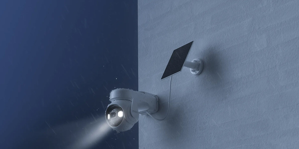 Na ścianie zamontowana jest zewnętrzna kamera z dwoma świecącymi diodami oraz panel solarny. Kamera jest skierowana w stronę ciemnego otoczenia, a na obrazie widać padający deszcz.