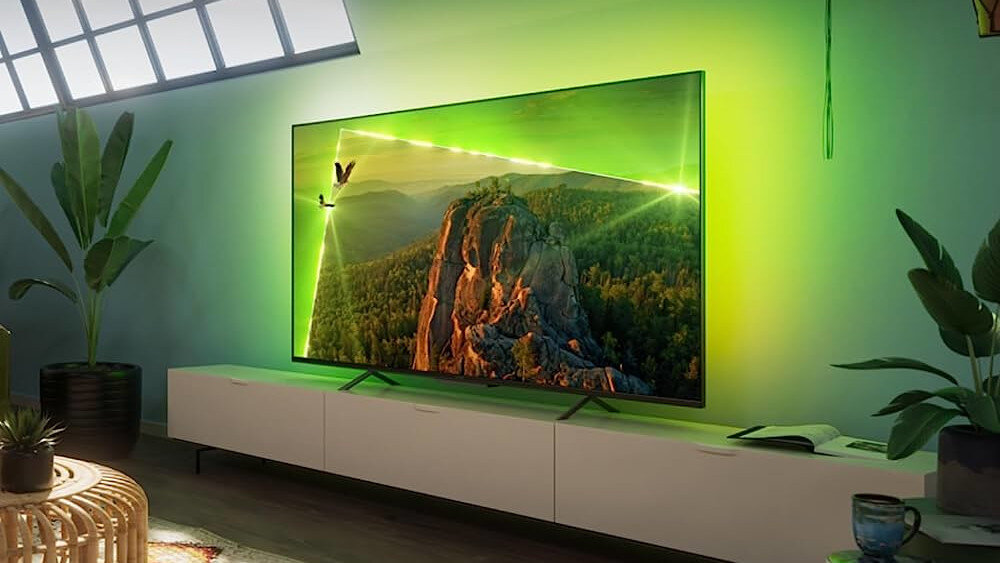 Telewizor wyświetla obraz górskiego krajobrazu z zielonym podświetleniem Ambilight na ścianie. Wnętrze pokoju jest nowoczesne, z białą szafką RTV i rośliną w doniczce.