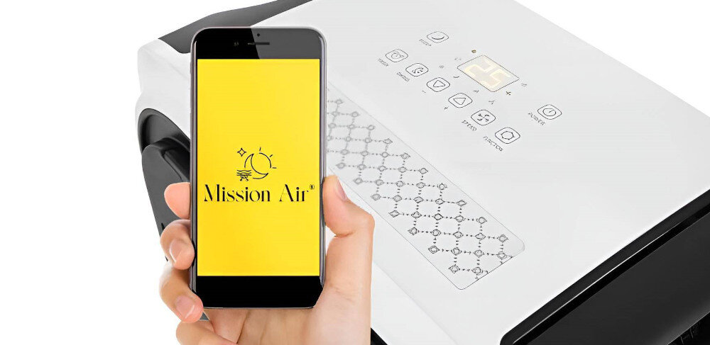 Osoba trzyma smartfon z żółtym ekranem, na którym widnieje napis \'Mission Air\'. W tle widoczny jest panel sterowania urządzenia z kilkoma przyciskami i wyświetlaczem.