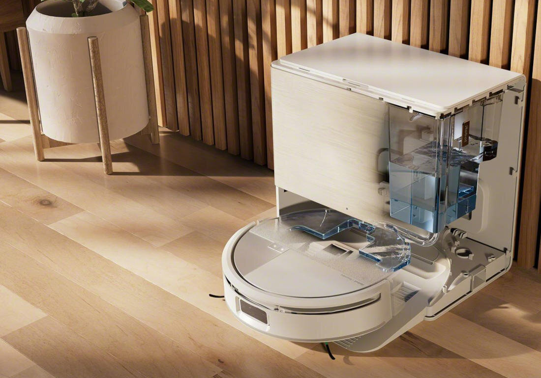 Na drewnianej podłodze stoi biały robot sprzątający z otwartą stacją dokującą. W tle widoczna jest drewniana ściana i doniczka z rośliną.