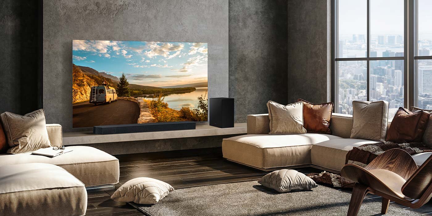 W eleganckim salonie znajduje się duży telewizor wyświetlający krajobraz z drogą i jeziorem, a przed nim umieszczony jest czarny soundbar. W pomieszczeniu są także jasne sofy, poduszki i duże okna z widokiem na miasto.
