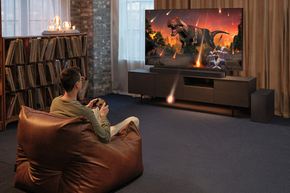Osoba siedzi na brązowym fotelu przed telewizorem, na którym wyświetlana jest scena z dinozaurem i spadającymi meteorytami. Obok telewizora znajduje się czarny soundbar i subwoofer.
