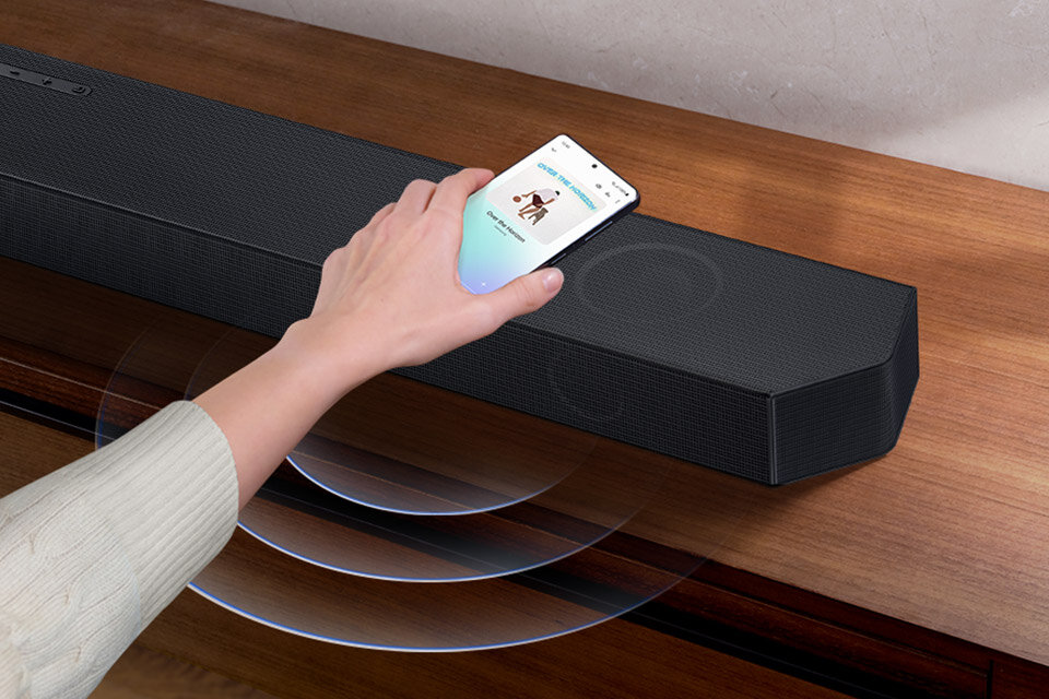 Czarny soundbar leży na drewnianej powierzchni, a nad nim znajduje się ręka trzymająca smartfon z wyświetlonym ekranem. Wokół soundbara widoczne są fale dźwiękowe.