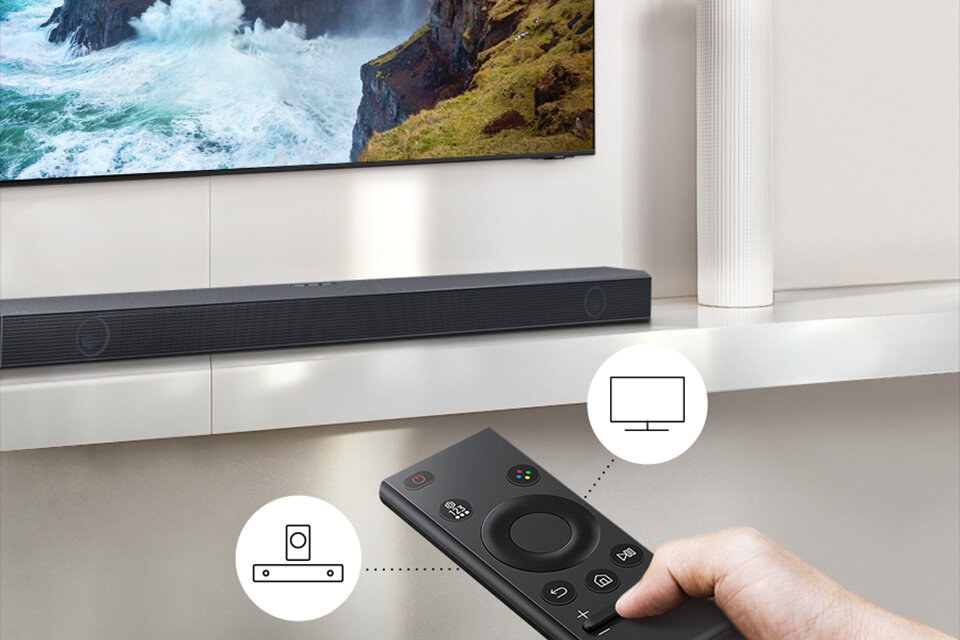 Na obrazie widoczny jest czarny soundbar umieszczony pod telewizorem, na którym wyświetlany jest krajobraz z morzem i klifami. W dolnej części zdjęcia znajduje się ręka trzymająca pilot z przyciskami, a obok widoczne są ikony przedstawiające soundbar i ekran.