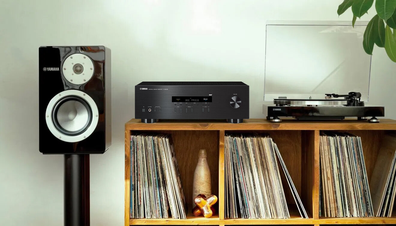 Na drewnianej półce znajduje się amplituner marki Yamaha oraz gramofon z przezroczystą pokrywą. Obok stoi głośnik z widocznym logo Yamaha, a poniżej półki widać kolekcję płyt winylowych.