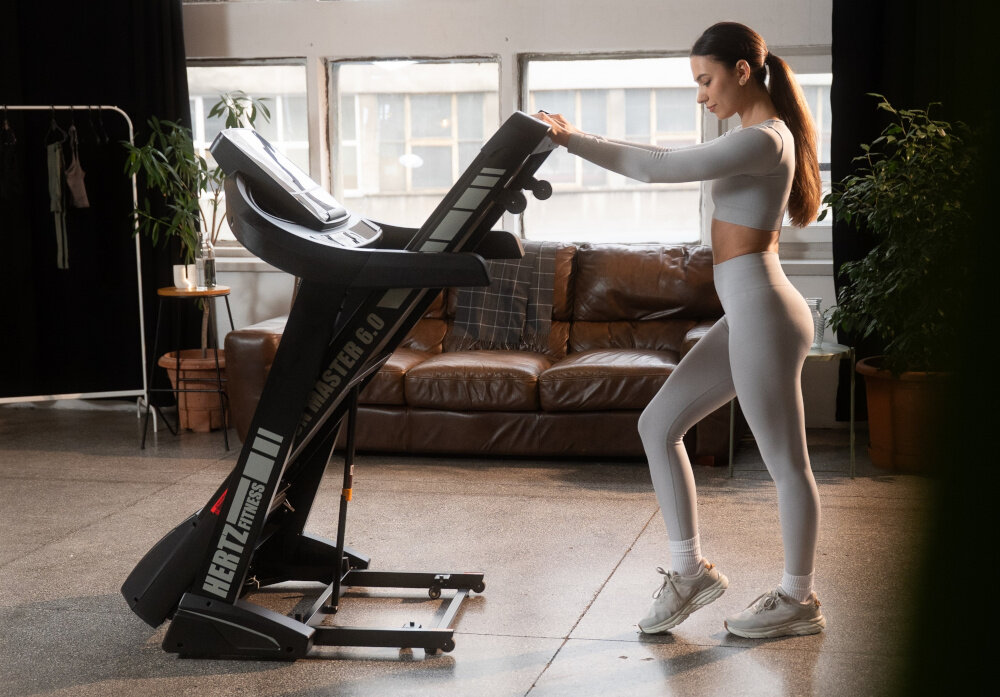 Kobieta w jasnym stroju sportowym stoi na bieżni elektrycznej HERTZ FITNESS Run Master 6.0. W tle znajduje się brązowa sofa i rośliny doniczkowe.
