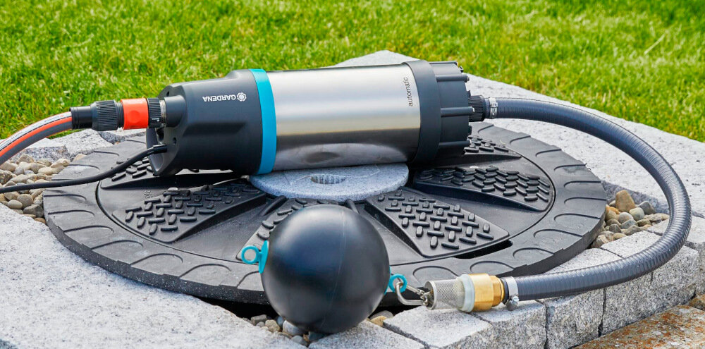 Na obrazie widoczna jest elektryczna pompa do wody z logo GARDENA, umieszczona na okrągłej podstawie z kamieniami i trawą w tle. Pompa jest podłączona do węża i ma metaliczną obudowę z niebieskim akcentem.