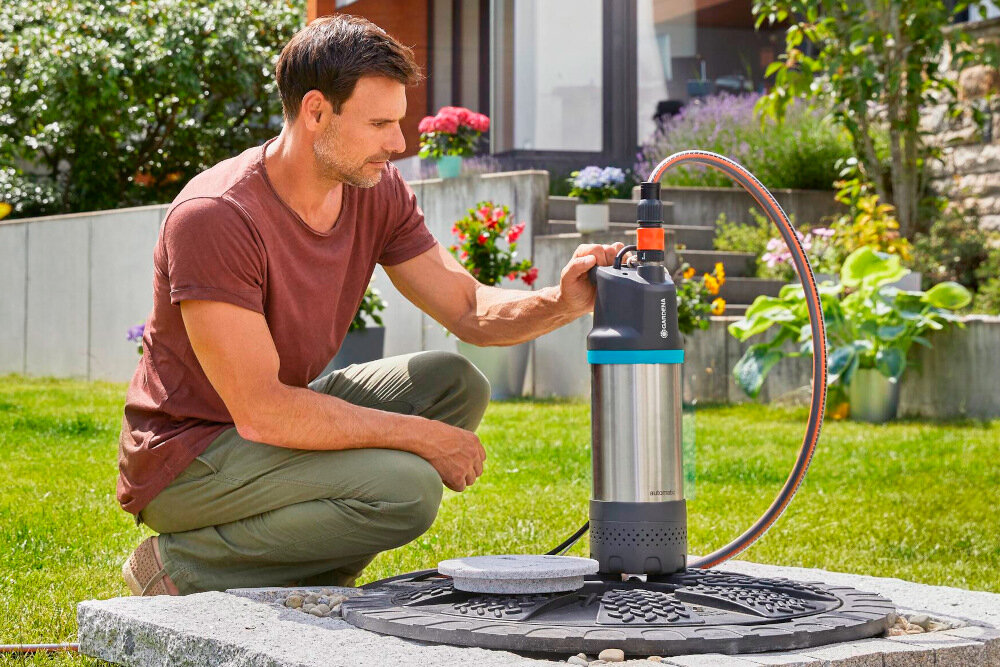 Osoba obsługuje elektryczną pompę do wody GARDENA na trawniku w ogrodzie. W tle widoczne są kwiaty i elementy architektury ogrodowej.