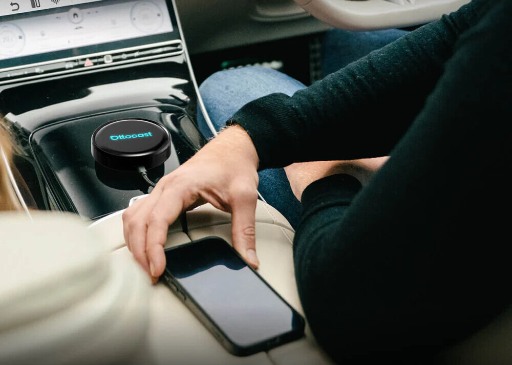 Na konsoli samochodowej znajduje się czarny adapter z logo \'Ottocast\'. Obok leży smartfon, a ręka osoby dotyka jego ekranu.
