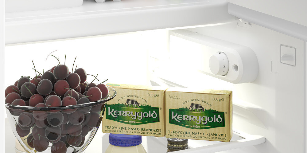 Wnętrze lodówki z widocznym pokrętłem temperatury, miską pełną ciemnych winogron oraz dwoma opakowaniami masła Kerrygold i słoikiem z niebieską zakrętką.