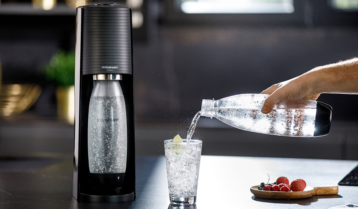 Na zdjęciu widać czarny saturator z logo \'sodastream\' oraz rękę nalewającą wodę z plastikowej butelki do szklanki z lodem i plasterkiem cytryny. Obok szklanki leży deska z owocami.