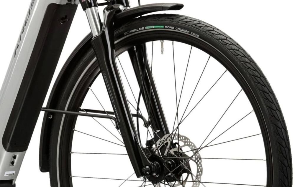 Zbliżenie na przednie koło roweru elektrycznego z widocznym amortyzatorem i tarczą hamulcową. Na oponie widoczny jest napis \'SCHWALBE ROAD CRUISER\'.