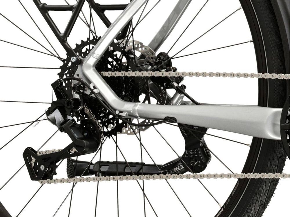 Zbliżenie na tylne koło roweru z widocznym mechanizmem przerzutki i łańcuchem. Rama roweru jest srebrna, a opona czarna.