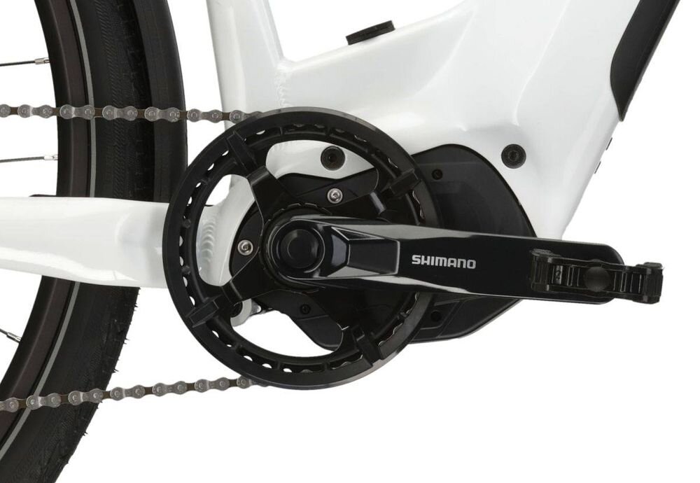 Zbliżenie na mechanizm korbowy roweru elektrycznego z widocznym napisem \'SHIMANO\' na ramieniu korby. Rama roweru jest w kolorze biało-srebrnym.