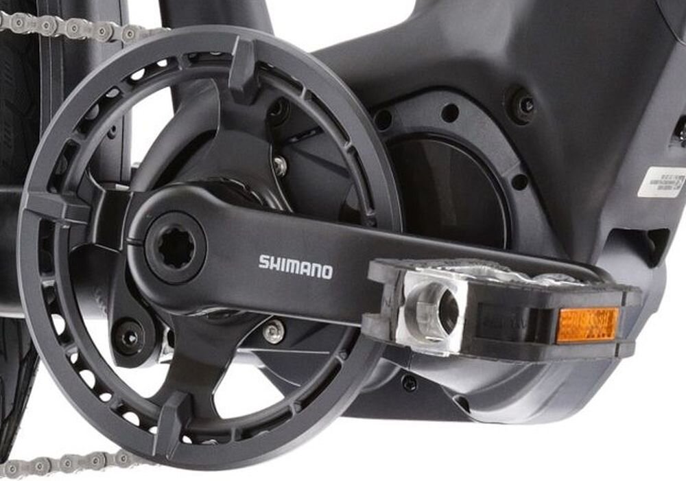 Zbliżenie na mechanizm korbowy roweru z widocznym napisem \'SHIMANO\' na ramieniu korby. Obok znajduje się pedał z odblaskiem.