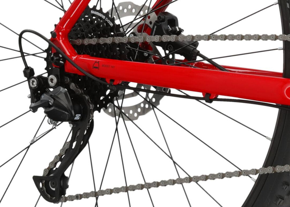 Zbliżenie na tylną część roweru górskiego z widocznym czerwonym ramieniem, mechanizmem przerzutki i łańcuchem. Na ramieniu znajduje się napis \'SHIMANO DEORE\'.