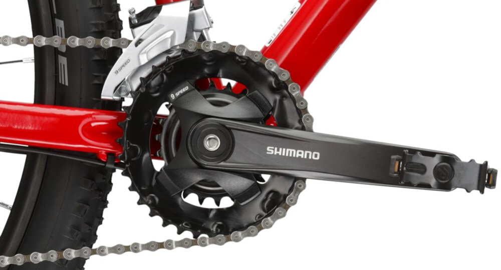Zbliżenie na mechanizm korbowy roweru z widocznym napisem \'SHIMANO\' na ramieniu korby. Rama roweru jest w kolorze czerwonym, a łańcuch jest zamontowany na zębatce.