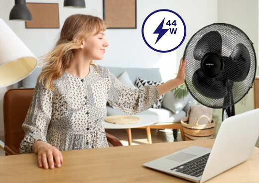 Kobieta siedzi przy biurku w pokoju. Obok niej stoi wentylator stojący MWP-15/C.