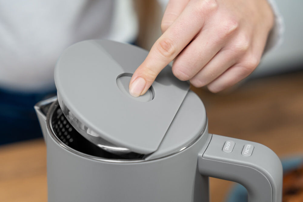 Osoba otwiera szary czajnik elektryczny, naciskając przycisk na pokrywie. Na uchwycie czajnika widoczne są dwa przyciski z napisami: \'ON/OFF\' i \'MENU\'.