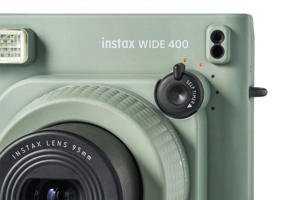 Zbliżenie na zielony aparat natychmiastowy z widocznym obiektywem i napisem \'instax WIDE 400\'. Obok obiektywu znajduje się pokrętło z napisem \'SELF TIMER\'.
