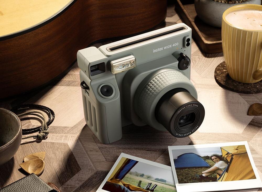 Na drewnianym stole znajduje się zielony aparat natychmiastowy z napisem \'instax WIDE 400\'. Obok aparatu leżą dwie wywołane fotografie, a w tle widoczna jest filiżanka z napojem i gitara.