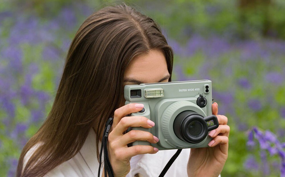 Osoba trzyma aparat natychmiastowy w kolorze zielonym z widocznym napisem \'instax wide 400\'. W tle znajduje się rozmyty krajobraz z fioletowymi kwiatami.