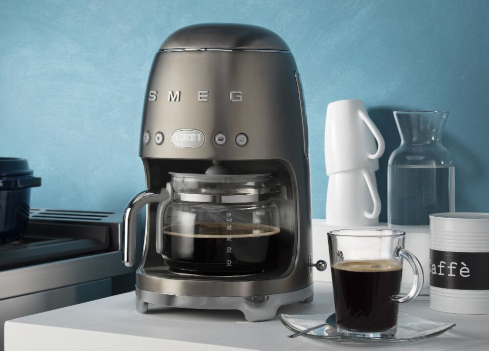 Na blacie stoi ekspres do kawy z napisem \'SMEG\', obok znajduje się szklanka z kawą. W tle widoczne są białe naczynia i pojemnik z napisem \'caffè\'.