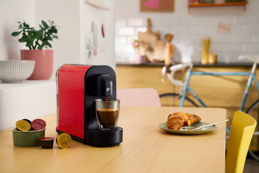 Na drewnianym stole stoi czerwony ekspres do kawy z filiżanką, obok znajdują się kapsułki do kawy. W tle widać kuchnię z rowerem i rośliną w doniczce.