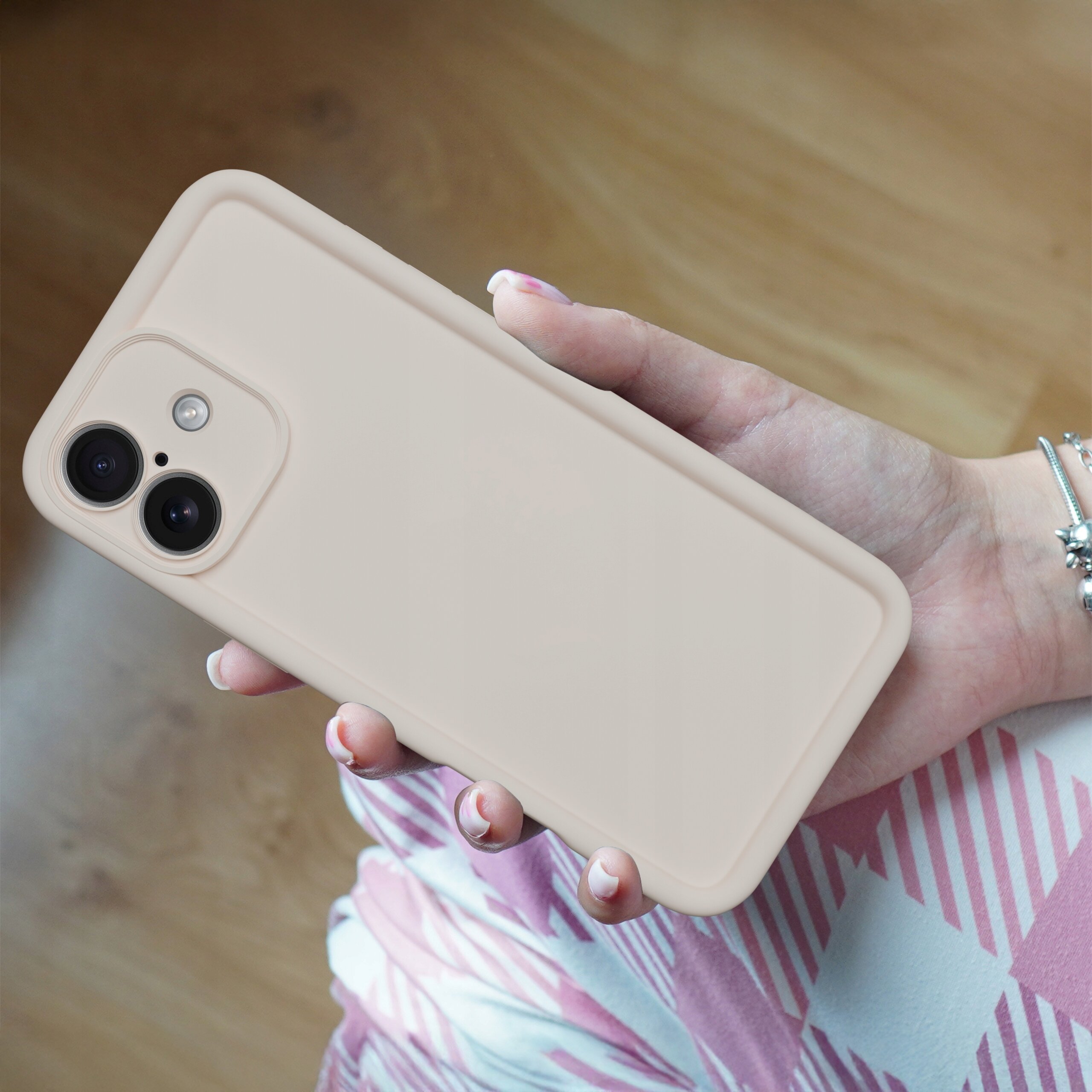 Osoba trzyma w dłoni beżowe, matowe etui na telefon z dwoma widocznymi aparatami fotograficznymi. W tle widać drewnianą podłogę i fragment ubrania w różowo-białe wzory.