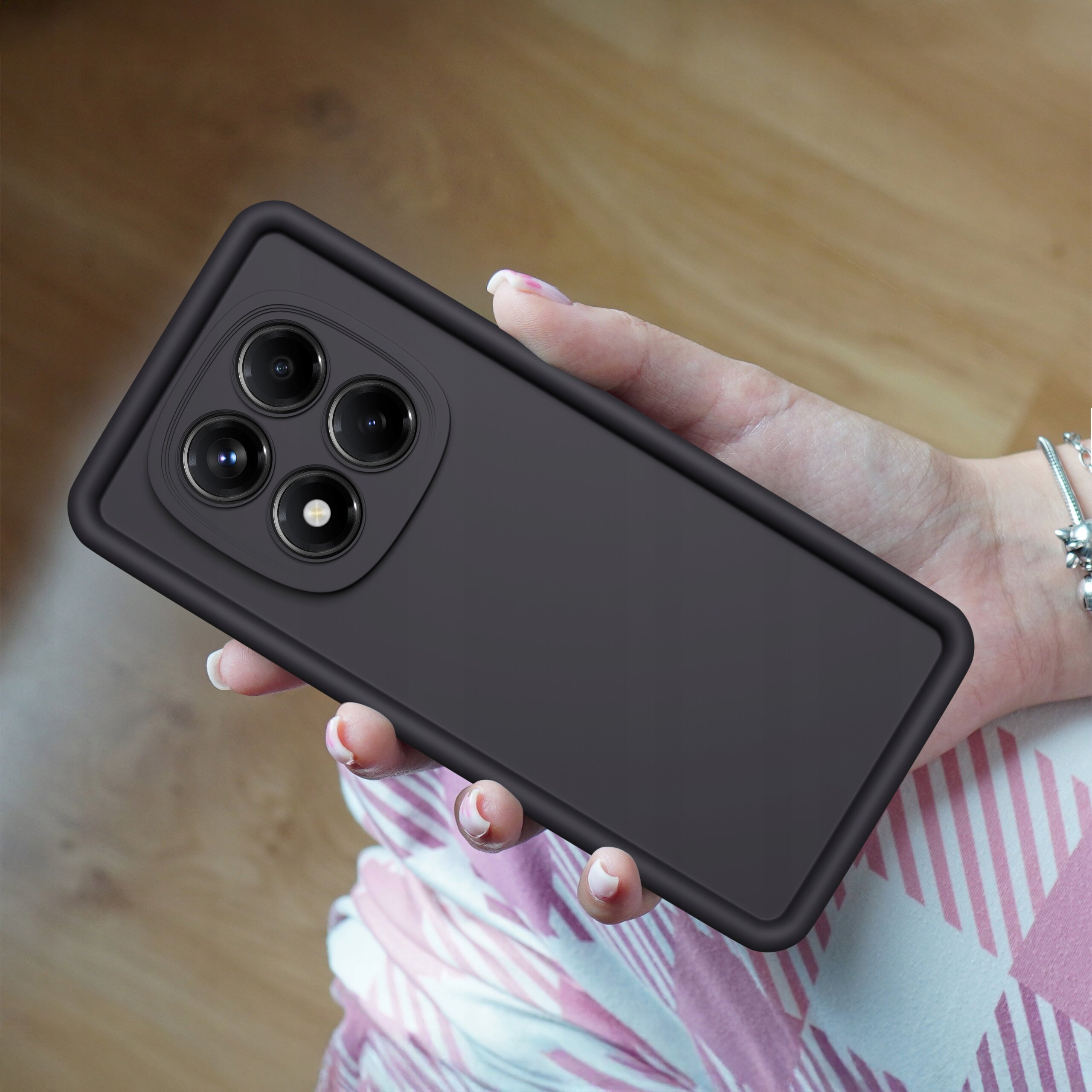 Osoba trzyma w dłoni czarne, matowe etui na telefon z trzema dużymi otworami na aparaty i jednym na lampę błyskową. Tło jest rozmyte, widać fragmenty ubrania w różowe i białe wzory.