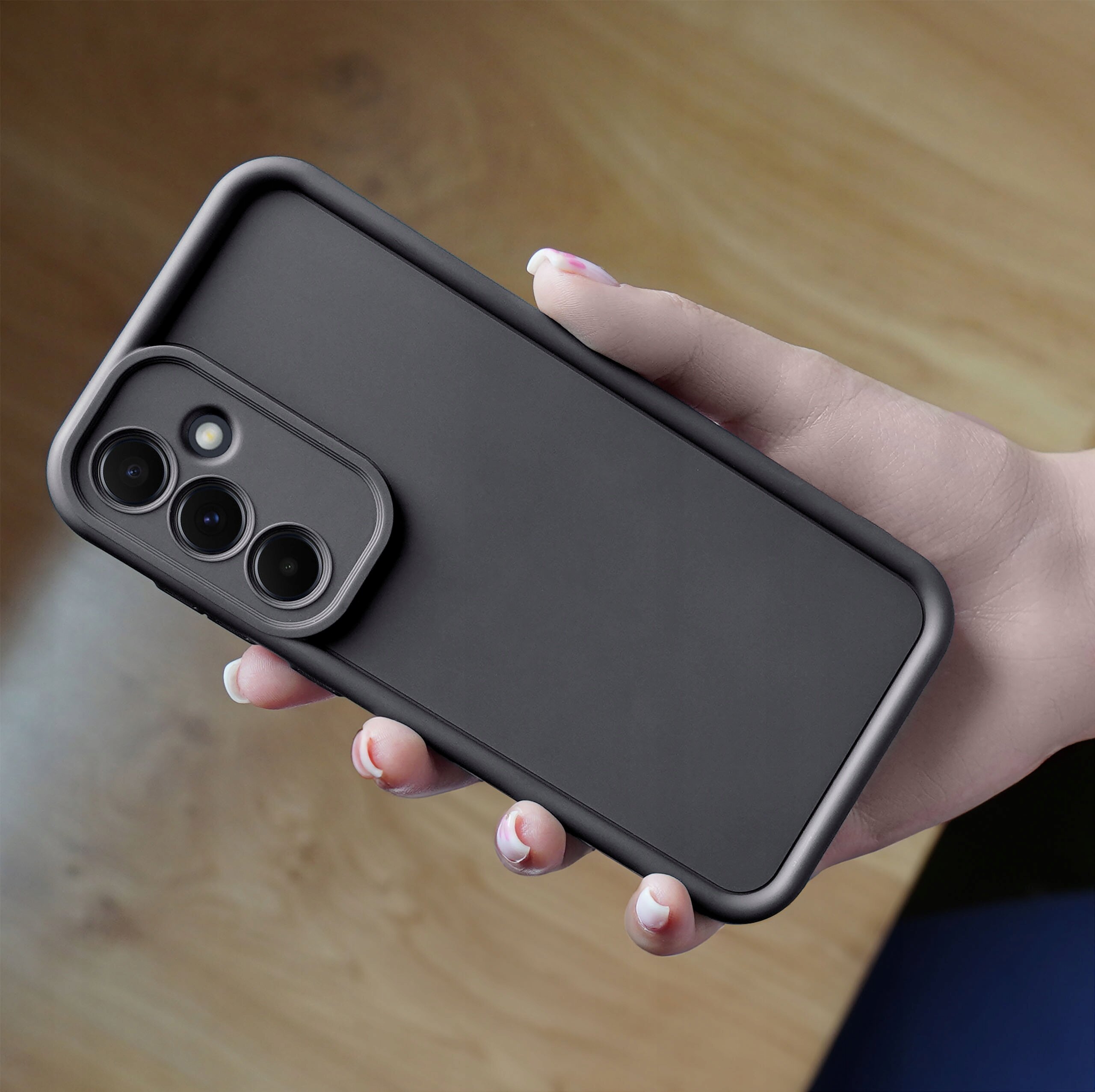 Czarny, matowy silikonowy etui na telefon z trzema otworami na aparaty fotograficzne, trzymane w dłoni na tle drewnianego blatu.