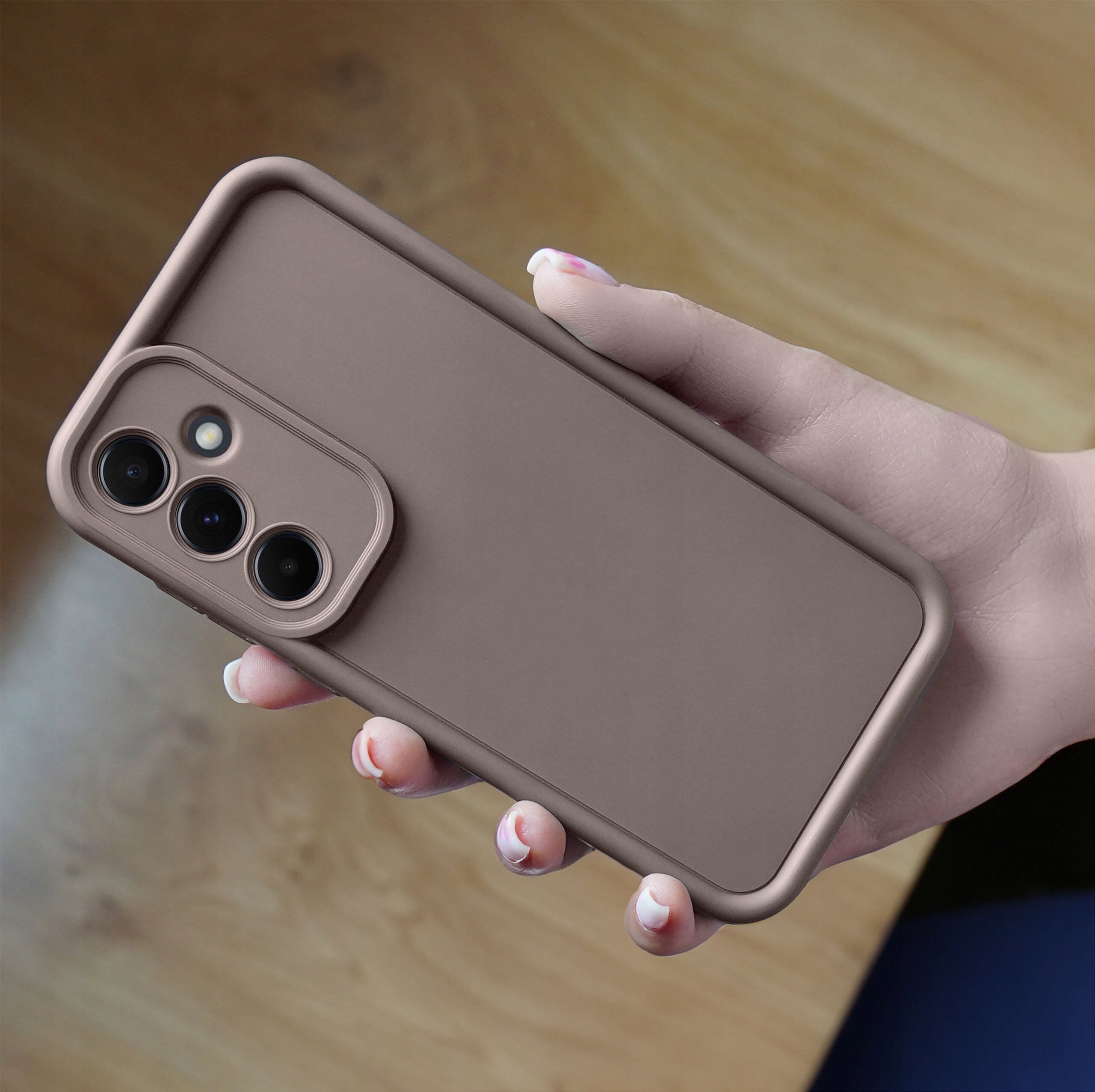 Brązowe, matowe etui silikonowe na telefon z trzema otworami na aparaty fotograficzne, trzymane w dłoni na tle drewnianej powierzchni.