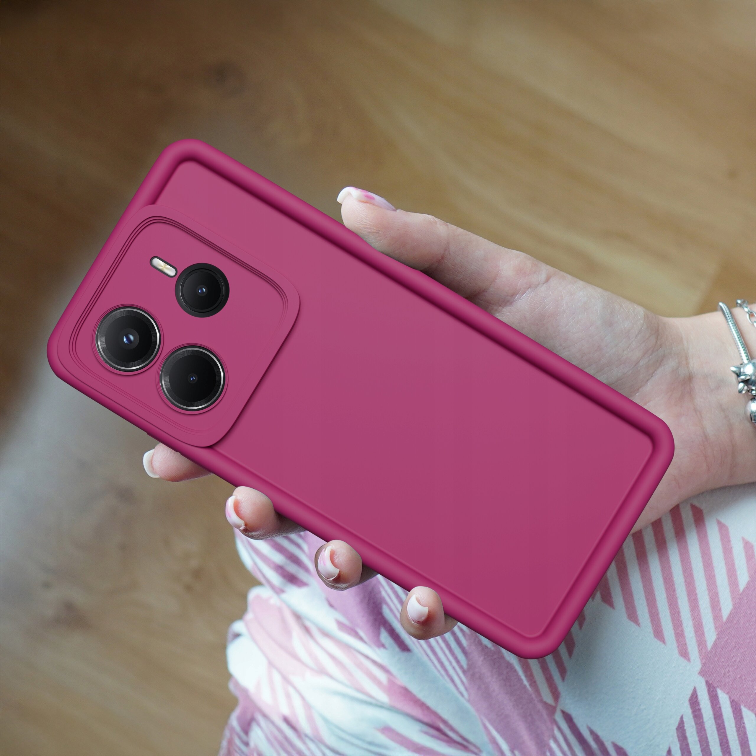 Osoba trzyma telefon w różowym, matowym etui z trzema widocznymi obiektywami aparatu. Tło jest drewniane, a na ubraniu widoczny jest wzór w różowe i białe paski.
