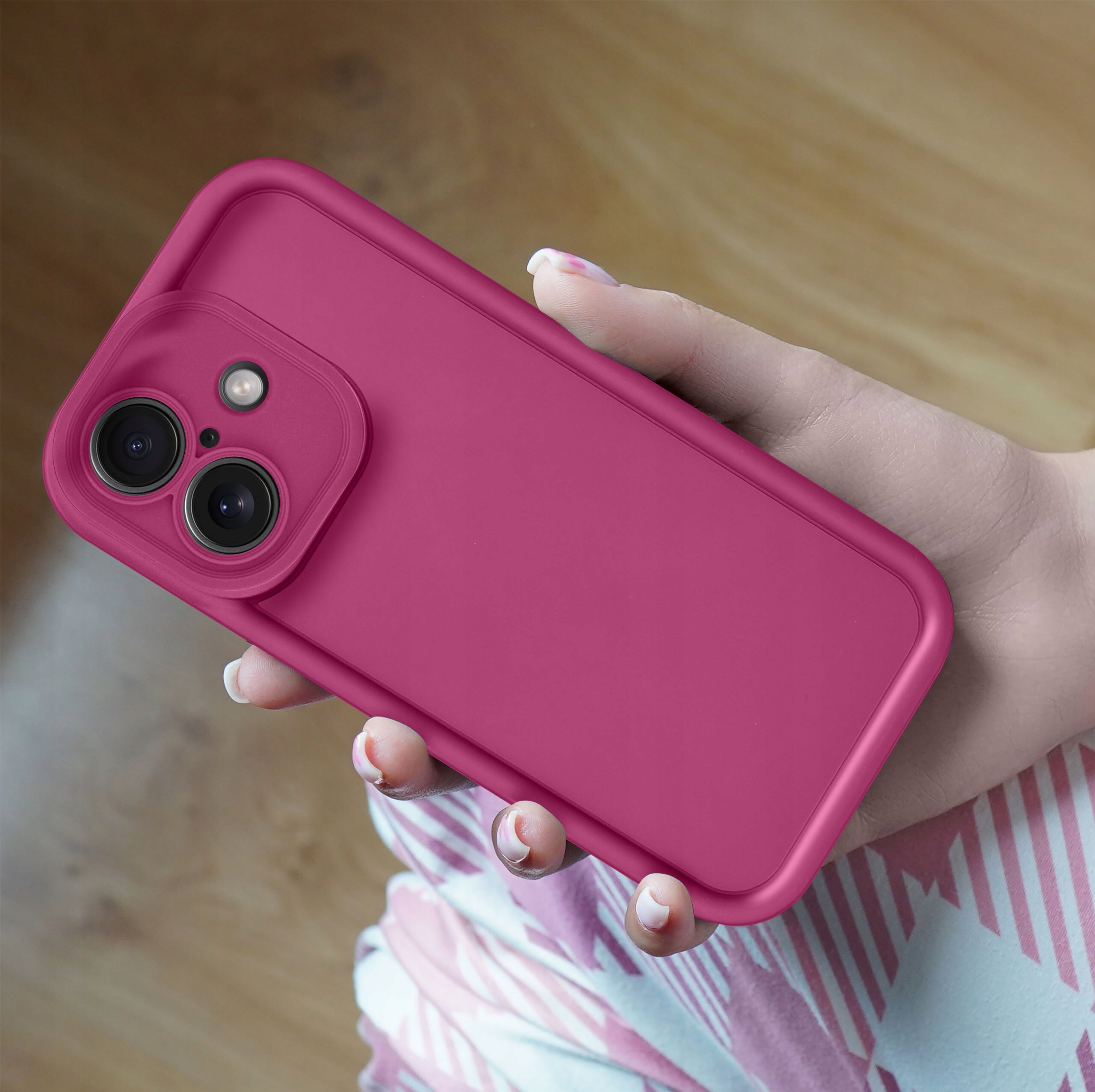 Różowe silikonowe etui na telefon z widocznymi dwoma aparatami fotograficznymi, trzymane w dłoni na tle drewnianej podłogi.