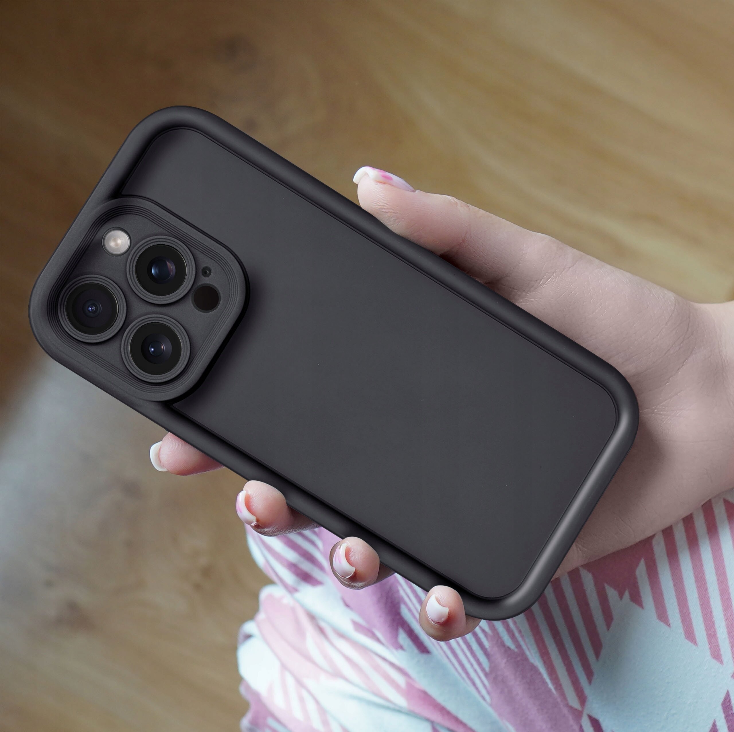 Osoba trzyma w dłoni telefon z czarnym, matowym etui, widoczne są trzy obiektywy aparatu i lampa błyskowa. Tło jest rozmyte, widać fragmenty ubrania w różowe i niebieskie wzory.