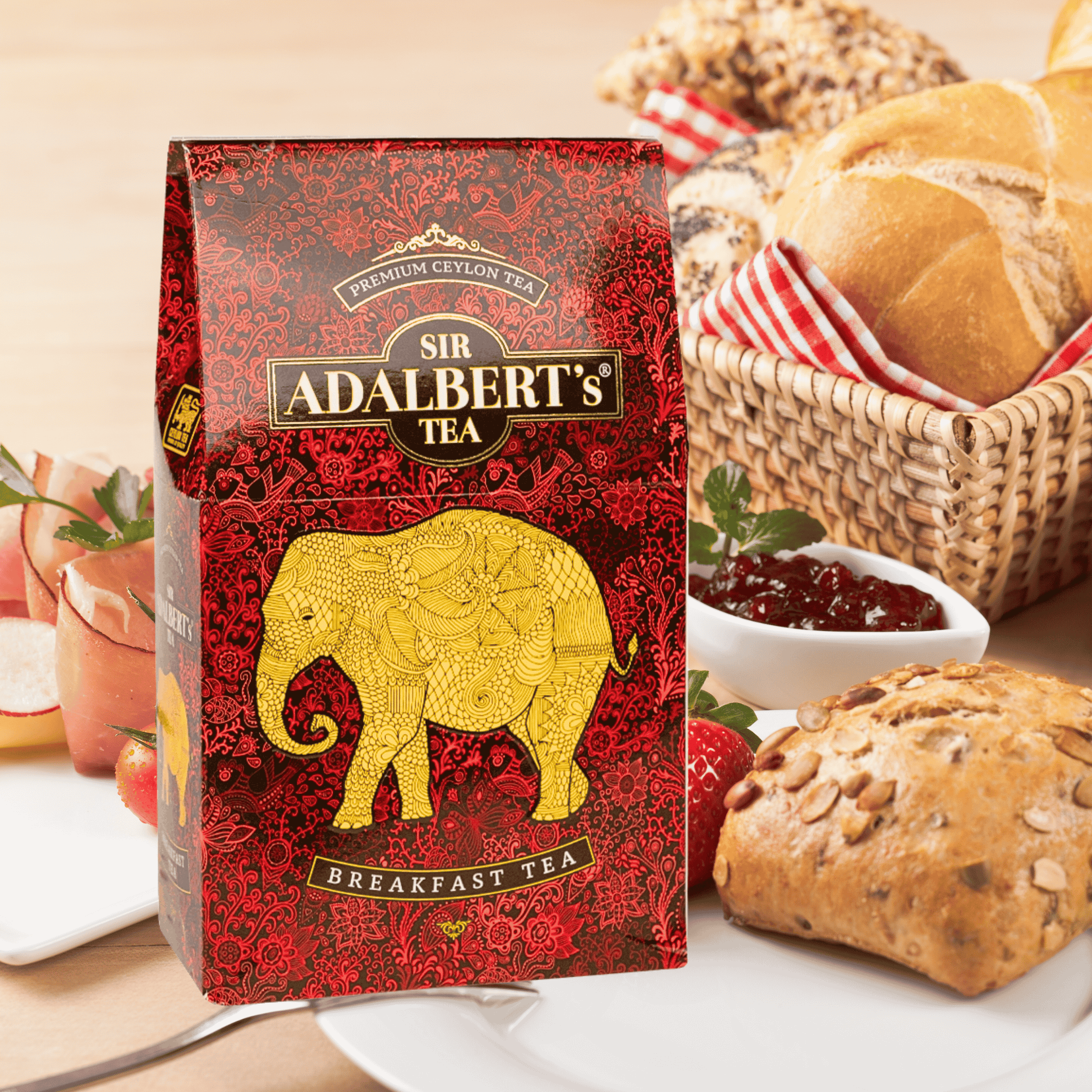 Opakowanie herbaty Sir Adalbert’s Breakfast Tea z czerwonym wzorem i złotym wizerunkiem słonia. W tle widoczne są pieczywo, dżem i owoce na stole.