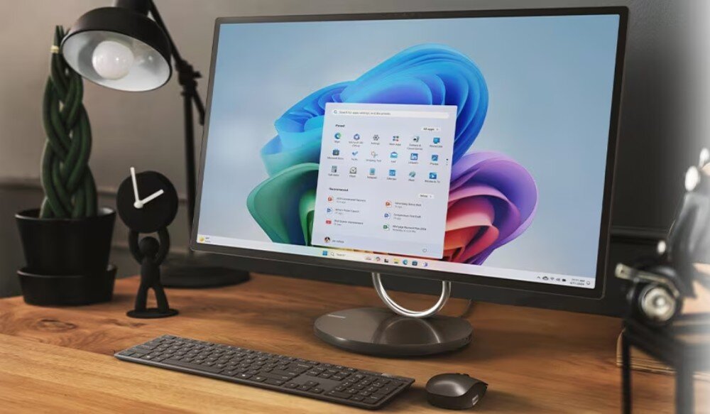 Na drewnianym biurku stoi komputer All in One z dużym ekranem, na którym wyświetlany jest pulpit systemu Windows 11. Obok znajduje się klawiatura, myszka oraz lampka biurkowa.