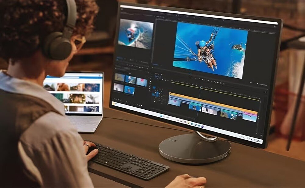 Osoba pracuje przy komputerze All in One z dużym monitorem, na którym widoczny jest program do edycji wideo. Obok znajduje się otwarty laptop z przeglądarką internetową wyświetlającą miniatury zdjęć.