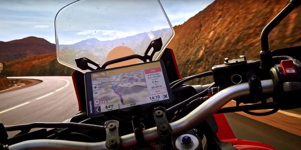 Motocykl z zamontowaną nawigacją GPS pokazującą mapę trasy i informacje o temperaturze oraz godzinie. W tle widoczna jest kręta droga i górzysty krajobraz.