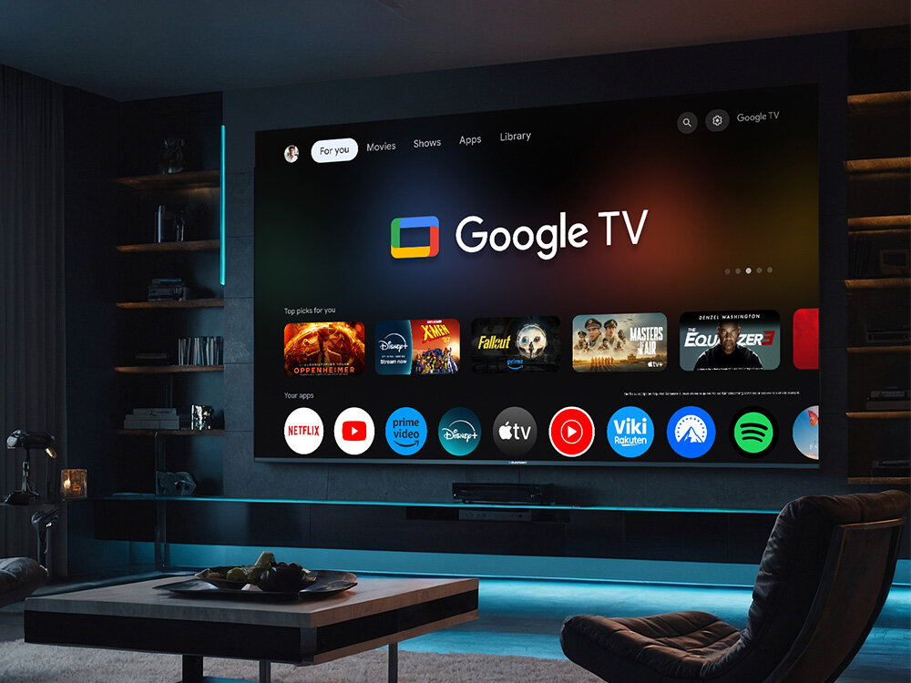 Duży ekran telewizora wyświetla interfejs Google TV z widocznymi ikonami aplikacji, takimi jak Netflix, Prime Video, Disney+, i Apple TV. W pomieszczeniu znajduje się nowoczesne wyposażenie, w tym półki z książkami i stolik kawowy.