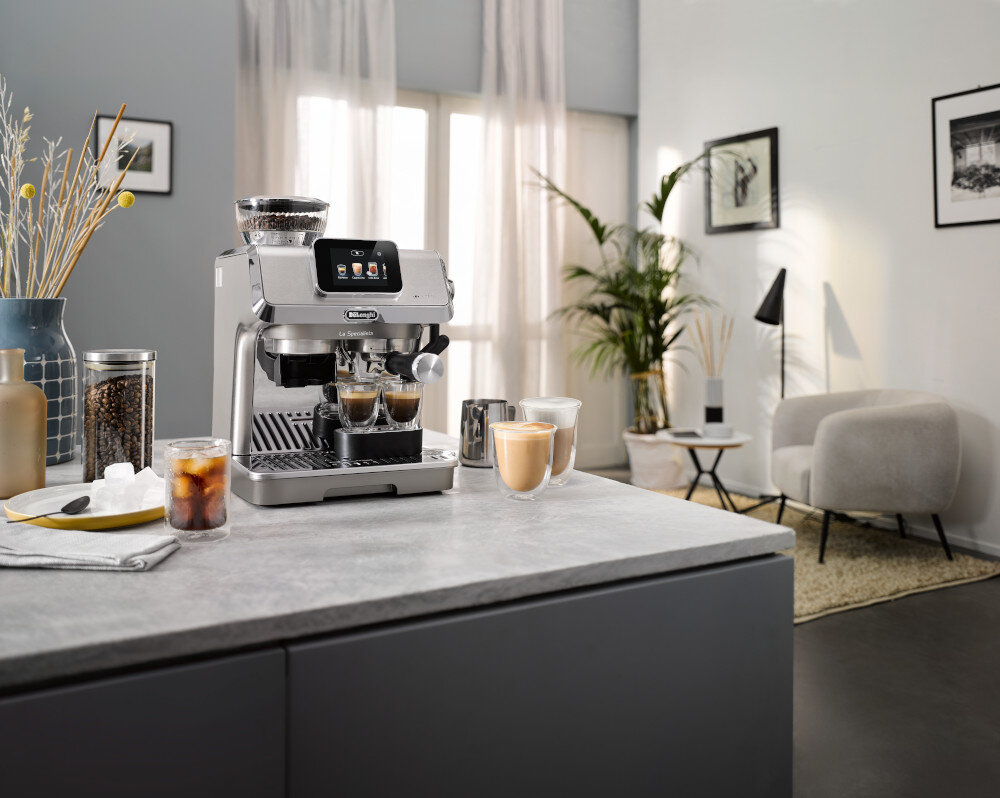 Na blacie kuchennym stoi ekspres do kawy z logo DELONGHI, obok którego znajdują się filiżanki z kawą. W tle widać jasne zasłony, roślinę doniczkową i nowoczesne meble.
