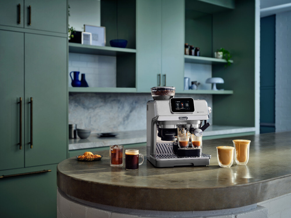 Na blacie kuchennym stoi srebrny ekspres do kawy z logo \'DeLonghi\', obok którego znajdują się filiżanki z kawą. W tle widoczne są zielone szafki kuchenne i półki z różnymi przedmiotami.