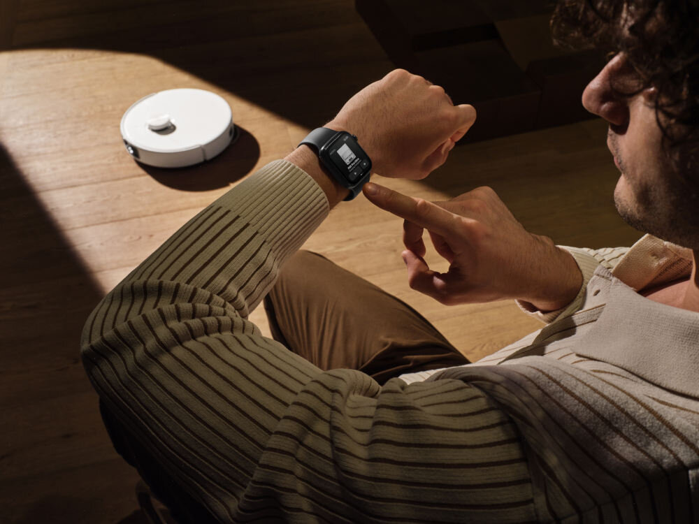 Osoba siedzi na podłodze, patrząc na smartwatch na nadgarstku, podczas gdy w tle widoczny jest biały robot sprzątający na drewnianej podłodze.