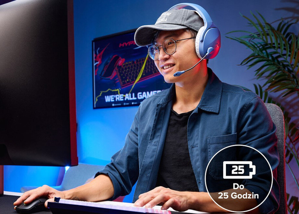 Osoba siedzi przy biurku, nosząc nauszne słuchawki z mikrofonem. W tle widoczny jest plakat z napisem \'WE\'RE ALL GAMERS\' oraz ikona baterii z tekstem \'Do 25 Godzin\'.
